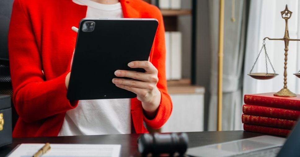 Mulher de blazer vermelho usa tablet em mesa de trabalho para automação rotina jurídica.